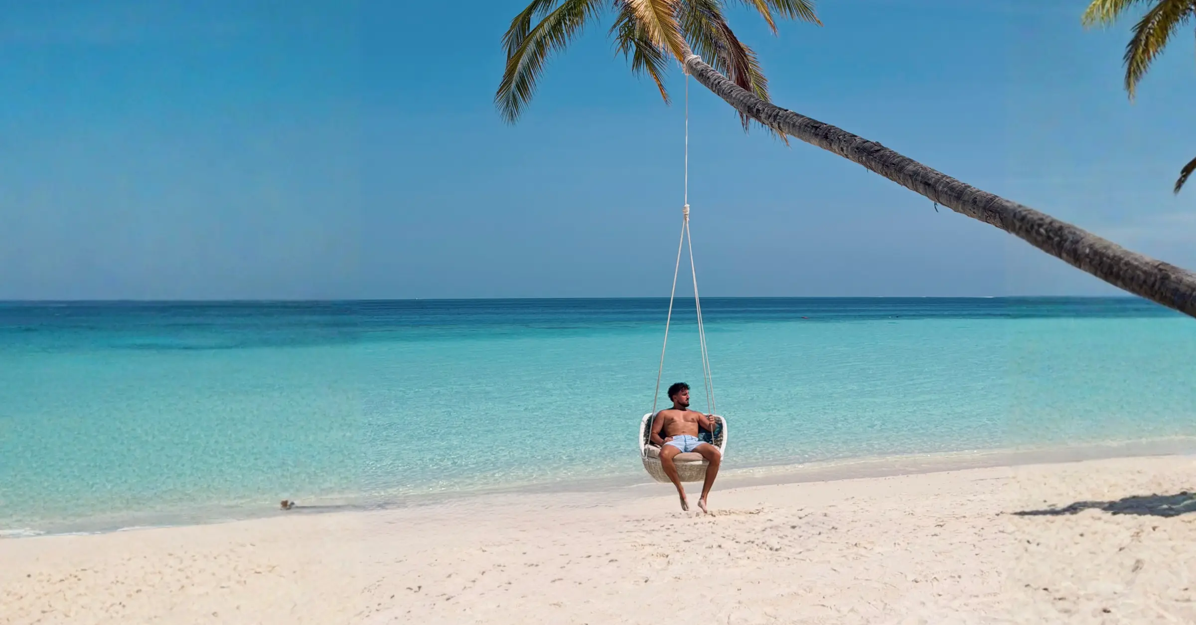 Mann sitzt in einer Hängeschaukel am weißen Sandstrand vor türkisblauem Meer