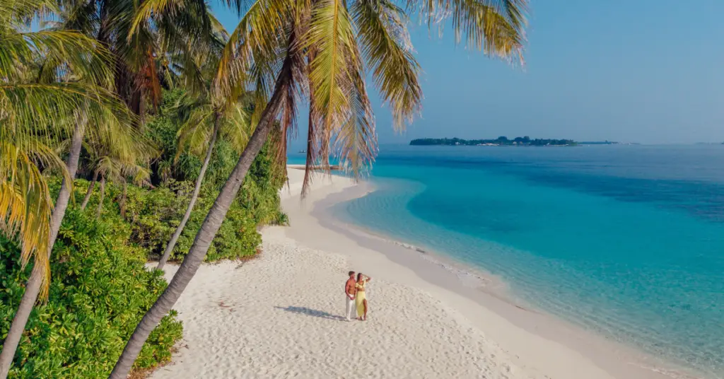 Palmen säumen einen weißen Sandstrand mit türkisblauem Meer; zwei Menschen stehen am Ufer