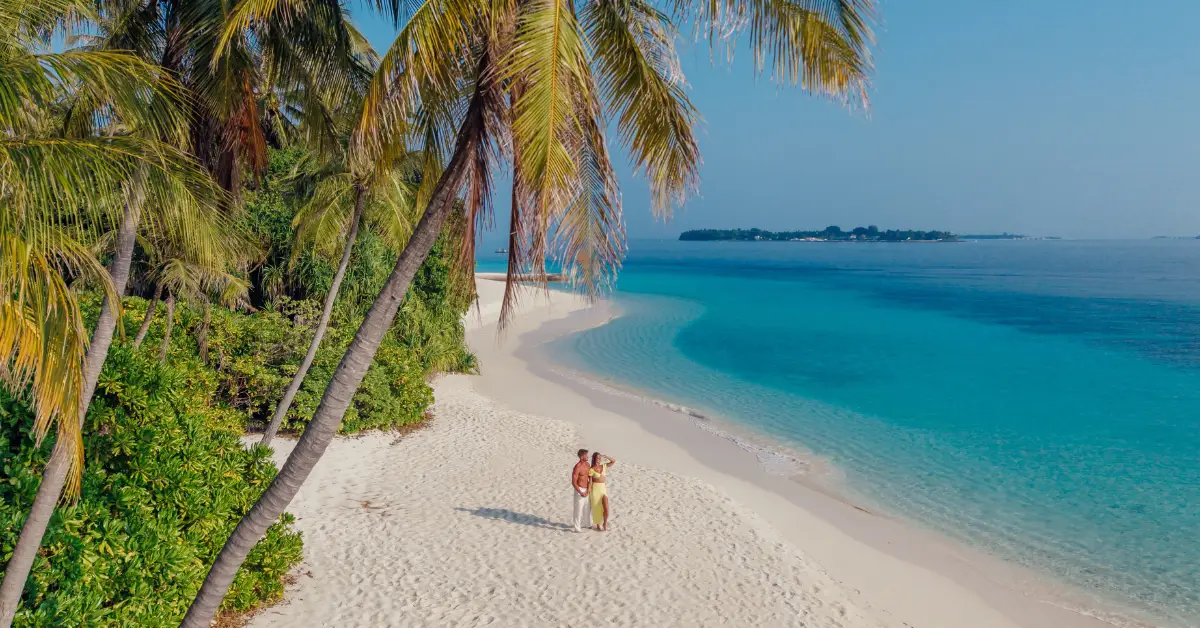 Palmen säumen einen weißen Sandstrand mit türkisblauem Meer; zwei Menschen stehen am Ufer