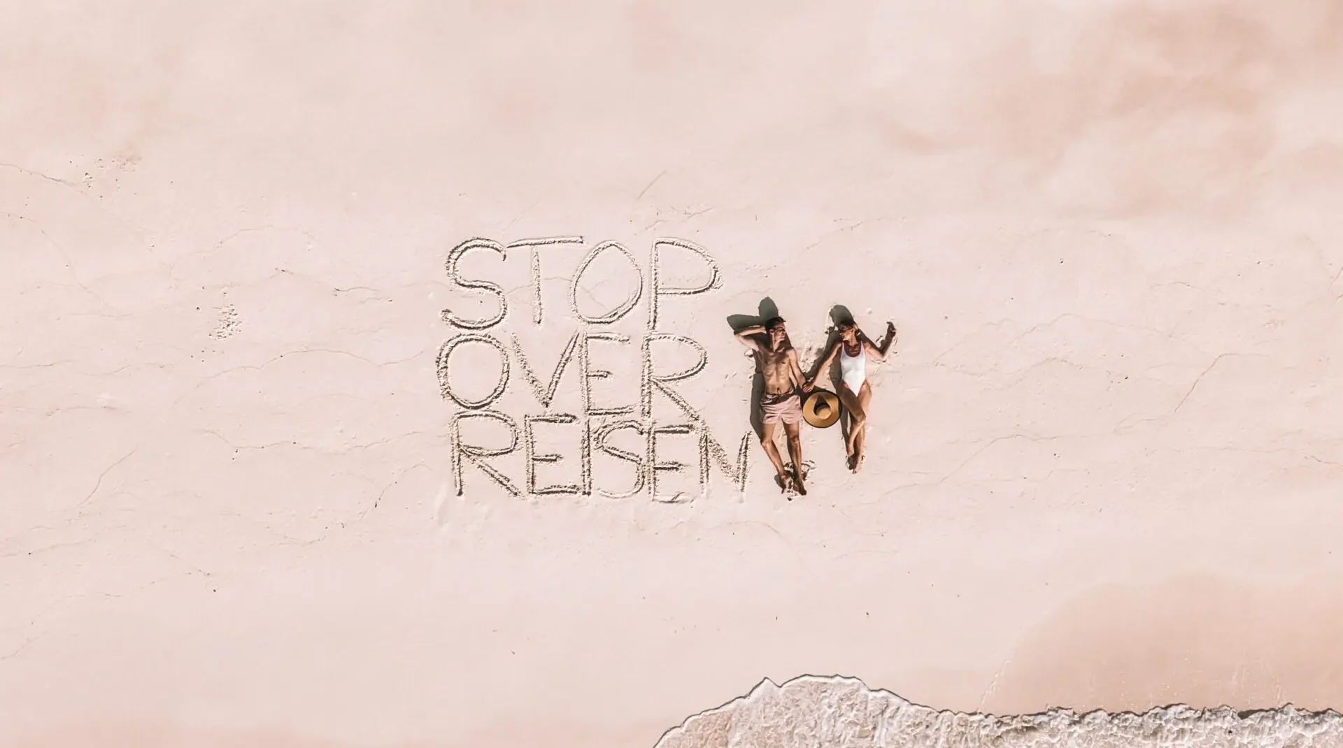Luftaufnahme eines Strandes mit den Worten „STOP OVER REISEM“ im Sand und zwei Personen daneben