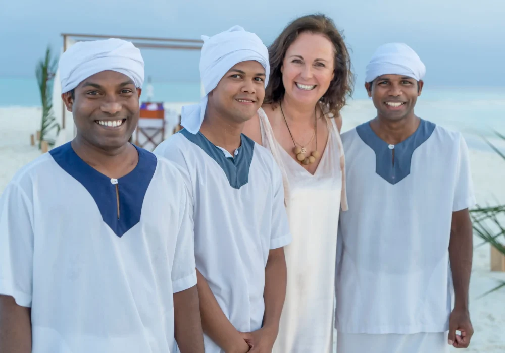 Drei Männer in traditioneller weißer Kleidung und eine lächelnde Frau stehen am Strand